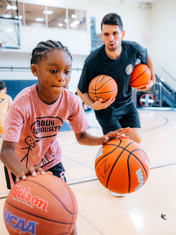 Carousel: a coach and a young player dribbling two basketballs at camp